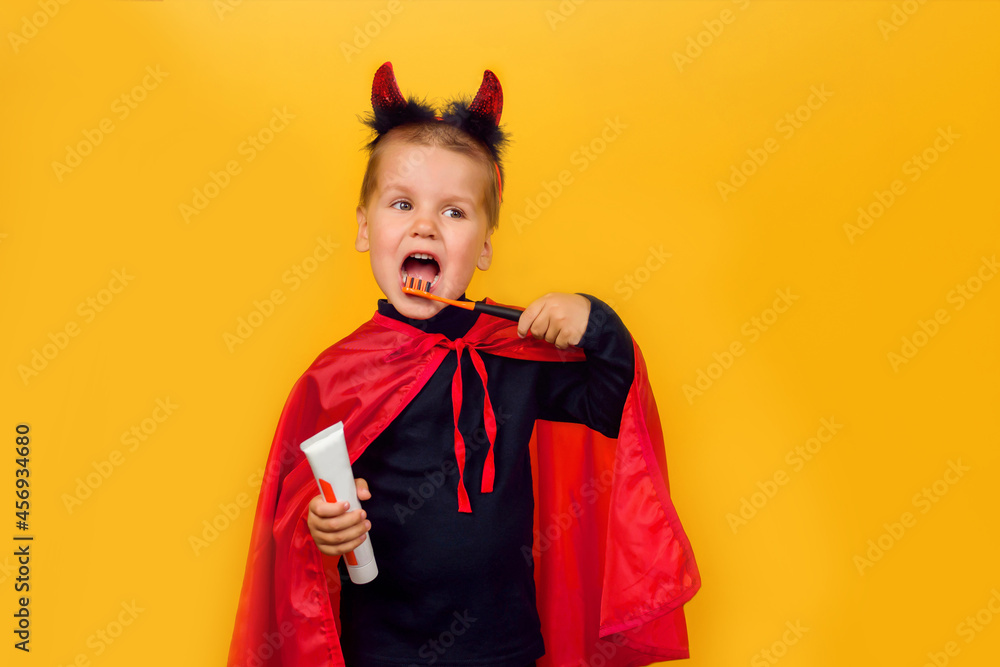 One little toddler boy in a carnival costume with toothpaste and an orange brush for Halloween is isolated on a yellow background. Medicine, dental hygiene, holidays concept. One little toddler boy in a carnival costume with toothpaste and an orange brush for Halloween is isolated on a yellow background. Medicine, dental hygiene, holidays concept.