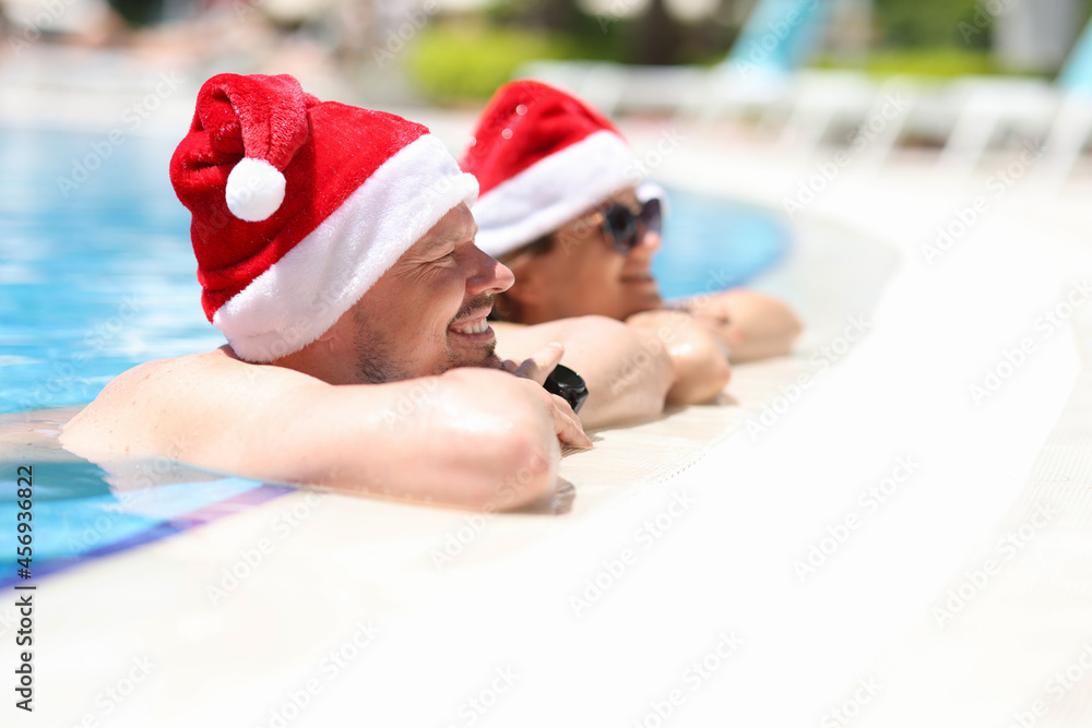 Fototapeta premium Young man and woman in sunglasses and santa claus hats lying on side of pool