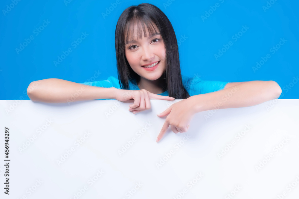 beautiful Asian young woman with bangs hair style in blue t shirt smiling and pointing finger a blank space for advertising banner , empty space white board a blank banner isolated on blue background