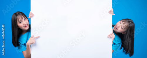 beautiful Asian woman with bangs hair style in blue t shirt smiling and pointing finger a blank space for advertising banner , empty copy space white board a blank banner isolated on blue background