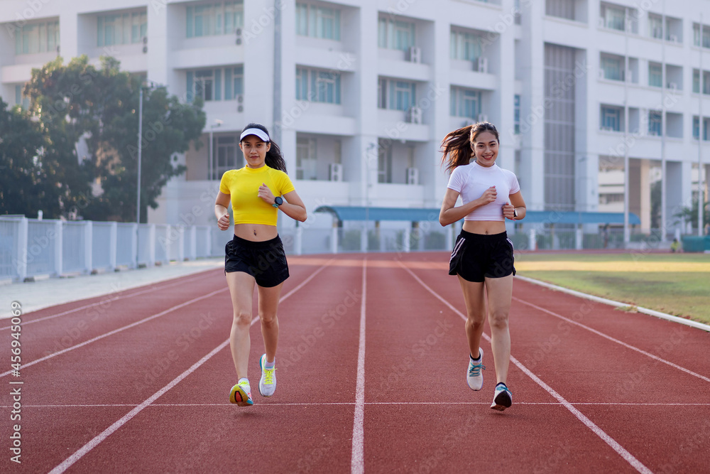 Two young Asian women in sports outfits jogging on running track in city stadium in the sunny morning for a healthy lifestyle. Young fitness women run on the stadium track.