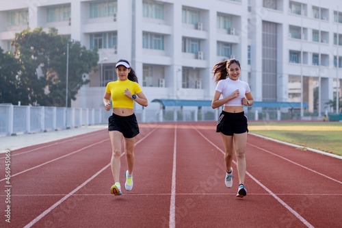 Two young Asian women in sports outfits jogging on running track in city stadium in the sunny morning for a healthy lifestyle. Young fitness women run on the stadium track.