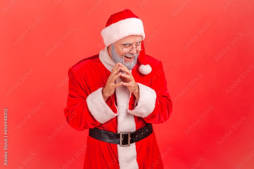 Insidious elderly man with gray beard wearing santa claus costume looking at camera with cunning tricky face and smirk, planning evil trick. Indoor studio shot isolated on red background. Insidious elderly man with gray beard wearing santa claus costume looking at camera with cunning tricky face and smirk, planning evil trick. Indoor studio shot isolated on red background.