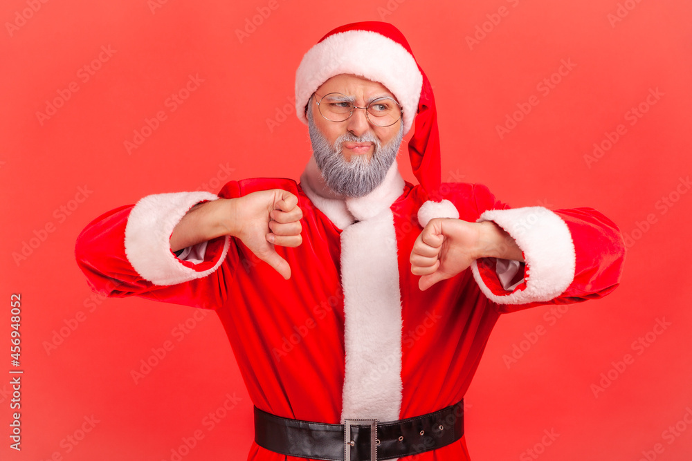 Elderly man with gray beard wearing santa claus costume has naughty expression and showing thumbs down, expressing disapproval, negative feedback. Indoor studio shot isolated on red background. Elderly man with gray beard wearing santa claus costume has naughty expression and showing thumbs down, expressing disapproval, negative feedback. Indoor studio shot isolated on red background.