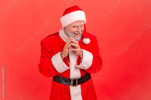 Insidious elderly man with gray beard wearing santa claus costume looking at camera with cunning tricky face and smirk, planning evil trick. Indoor studio shot isolated on red background.