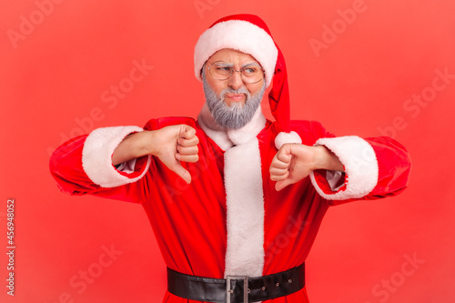 Фотография Elderly man with gray beard wearing santa claus costume has naughty expression and showing thumbs down, expressing disapproval, negative feedback