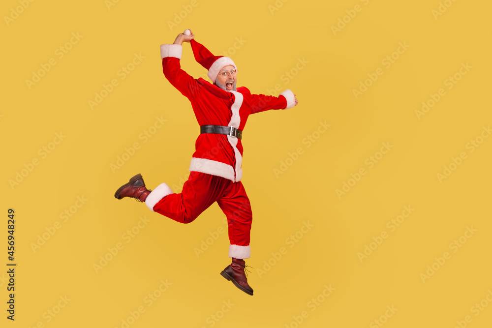 Full length of elderly man with gray beard wearing santa claus costume jumping up high and pulling his red hat, celebrating Christmas with excitement. Indoor studio shot isolated on yellow background.