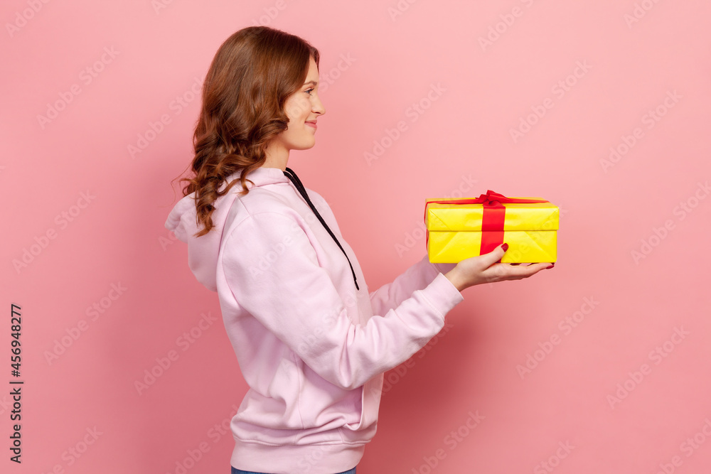Profile of cute curly haired teenage girl in casual hoodie holding wrapped gift box, holiday greeting, pleasant surprise for event. Indoor studio shot isolated on pink background Profile of cute curly haired teenage girl in casual hoodie holding wrapped gift box, holiday greeting, pleasant surprise for event. Indoor studio shot isolated on pink background