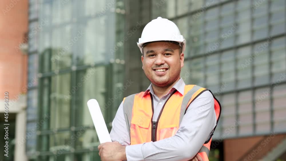 Asian engineer handsome man or architect looking forward holding paperwork blueprint with white safety helmet in construction site. Standing at modern building. Worker asian man with arm crossed Asian engineer handsome man or architect looking forward holding paperwork blueprint with white safety helmet in construction site. Standing at modern building. Worker asian man with arm crossed