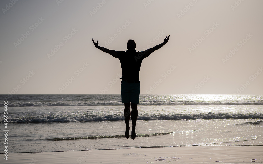 silhouette of athletic man jumping with raised hands on summer beach, freedom