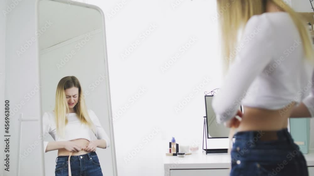 A young woman stands in front of a mirror and measures the circumference of her waist with a tape measure. The girl is complexed because of the weight A young woman stands in front of a mirror and measures the circumference of her waist with a tape measure. The girl is complexed because of the weight