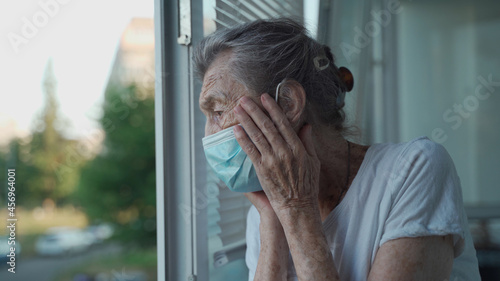 Worried senior woman in a protective medical mask sadly looks out the window with her head clasped in her hands in a nursing home. Depressed lady at home during the covid-19 pandemic