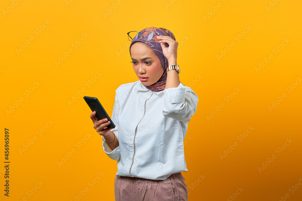 Shocked Asian woman looking at smartphone and holding glasses over yellow background Shocked Asian woman looking at smartphone and holding glasses over yellow background