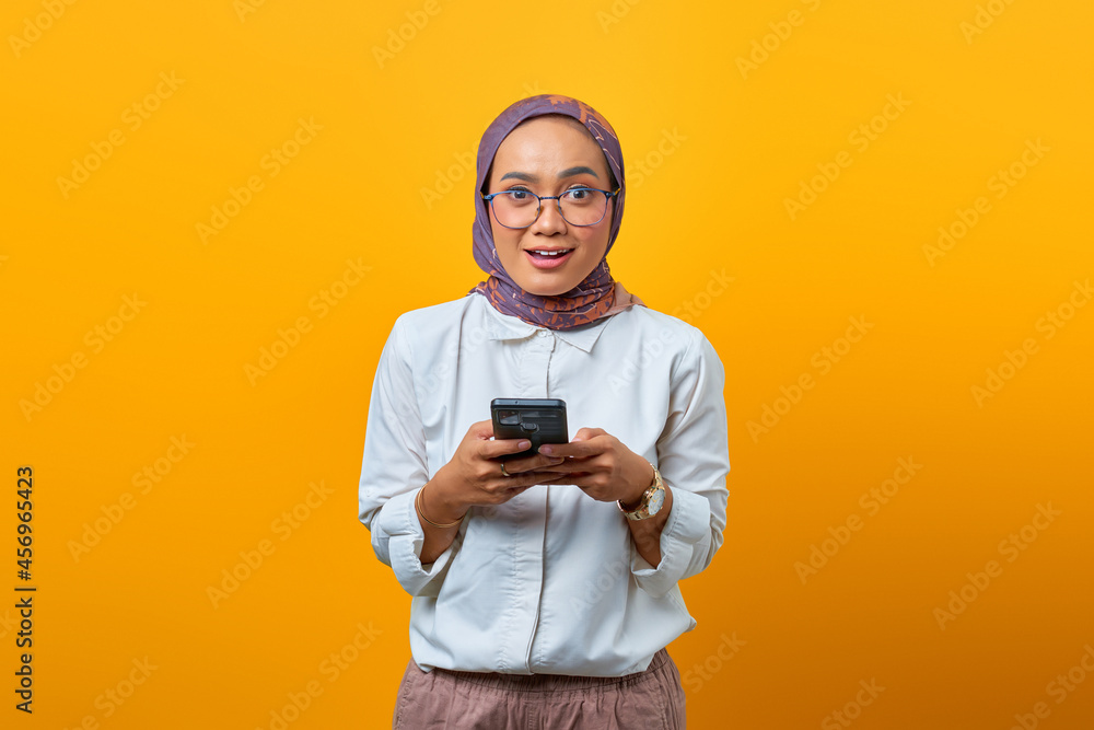 Shocked Asian woman using mobile phone and looking at camera over yellow background Shocked Asian woman using mobile phone and looking at camera over yellow background
