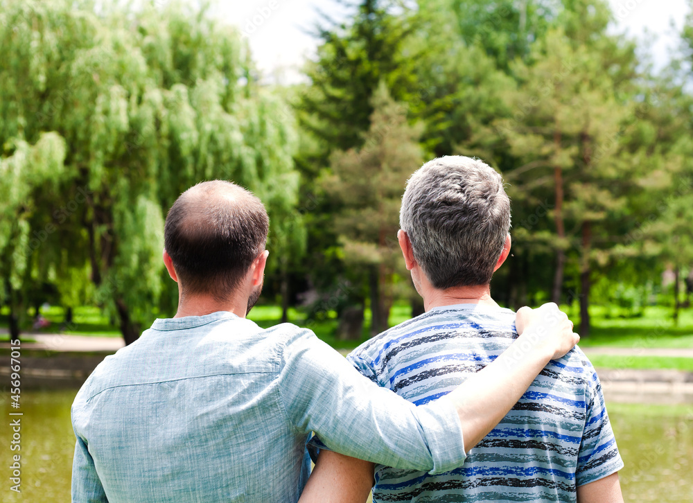 Adult father and son are hugging each other standing in the park. Back view. Adult father and son are hugging each other standing in the park. Back view.
