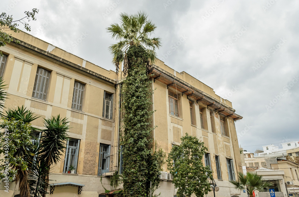 Large Neoclassic Building Facade in Downtown Heraclion, Crete Island, Greece with a High Palm Tree on a Cloudy Day.