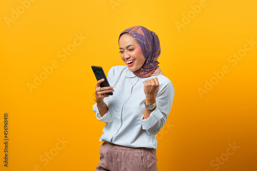 Excited Asian woman holding mobile phone celebrating luck over yellow background