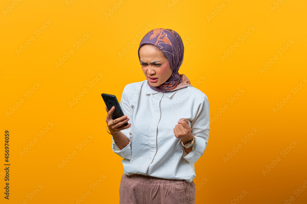 Angry Asian woman looking at smartphone getting bad news over yellow background Angry Asian woman looking at smartphone getting bad news over yellow background
