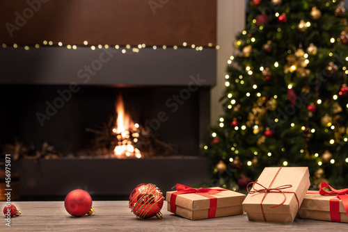 Cozy Christmas atmosphere with decorations and gifts on wooden table in the foreground. In the background blurred the fireplace with fire, the Christmas tree and Christmas decorations. Selective focus