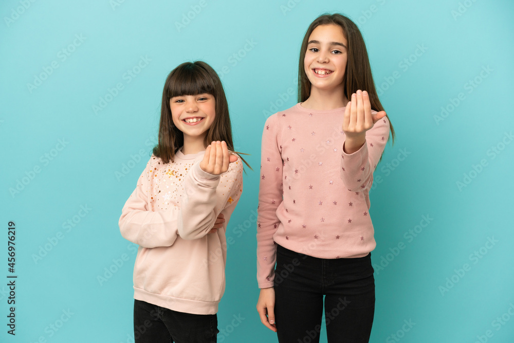 Little sisters girls isolated on blue background inviting to come with hand. Happy that you came