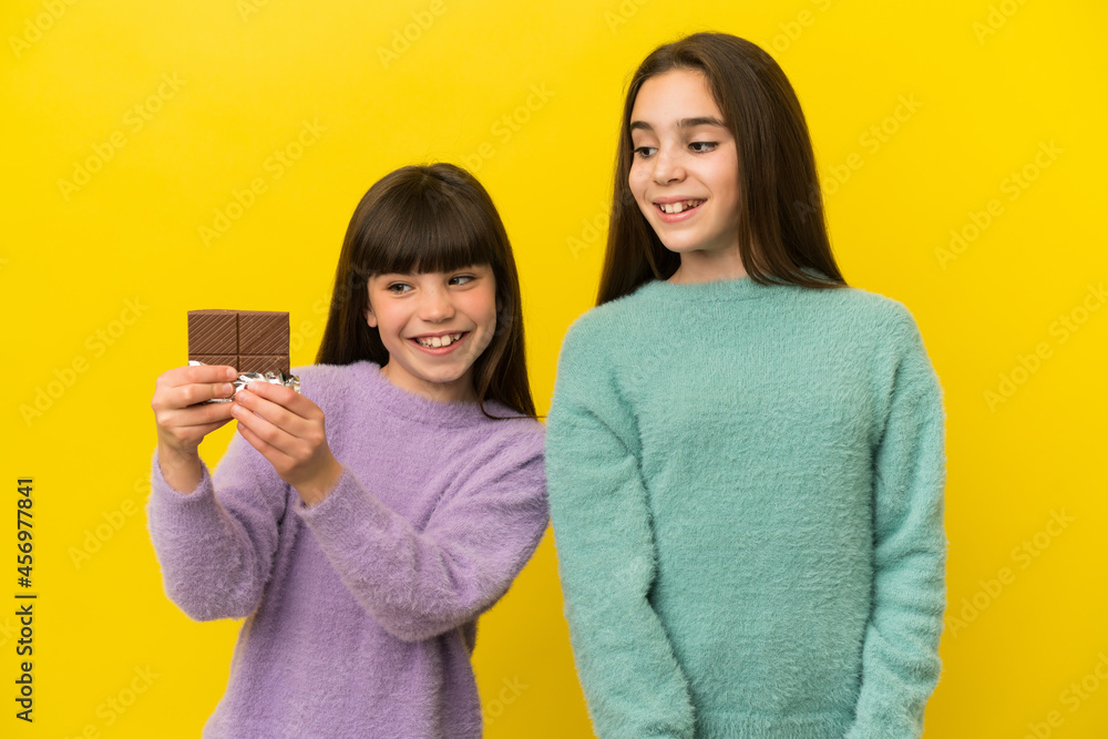 Little sisters isolated on yellow background taking a chocolate tablet and happy Little sisters isolated on yellow background taking a chocolate tablet and happy