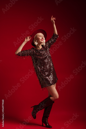 Happy smiling fashionable blonde woman wearing trendy marsala color sequin mini dress, hat, high leather boots, dancing, posing, on dark red background. Full length studio portrait