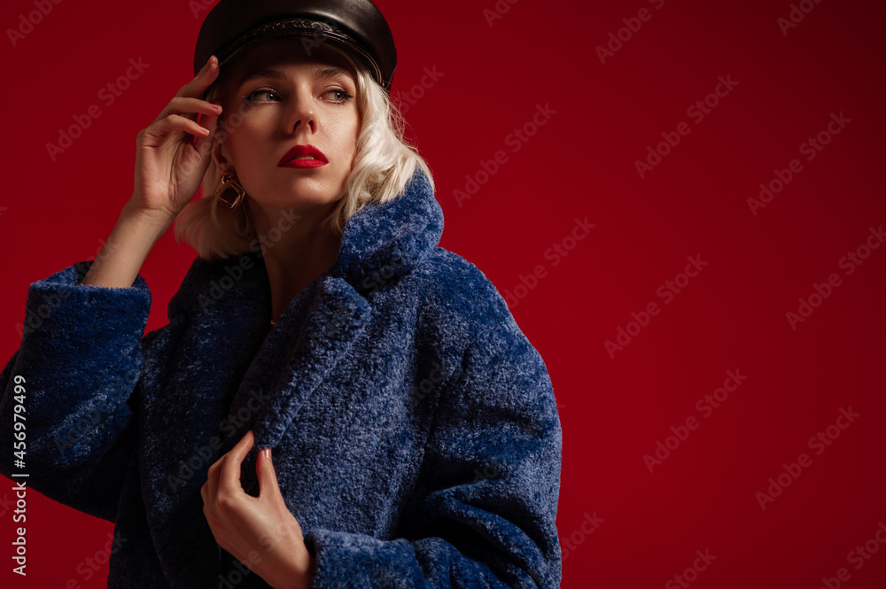 Fashion portrait of elegant confident blonde woman with red lips makeup, wearing trendy leather cap, blue faux fur coat, posing on dark red background. Copy, empty space for text Fashion portrait of elegant confident blonde woman with red lips makeup, wearing trendy leather cap, blue faux fur coat, posing on dark red background. Copy, empty space for text