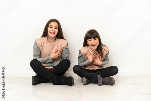 Little sisters sitting on the floor isolated on white background smiling a lot while putting hands on chest