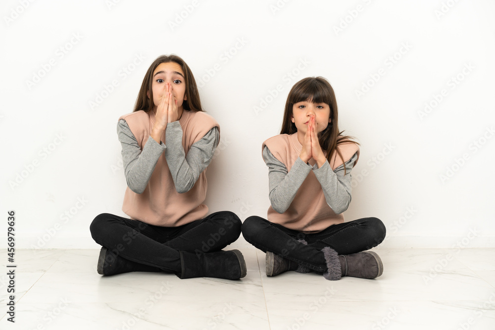 Little sisters sitting on the floor isolated on white background keeps palm together. Person asks for something Little sisters sitting on the floor isolated on white background keeps palm together. Person asks for something
