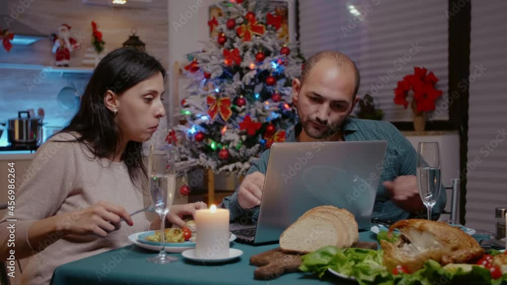 Festive couple looking at laptop and enjoying christmas dinner with technology. People eating chicken and drinking champagne while using device. Man and woman celebrating holiday. Festive couple looking at laptop and enjoying christmas dinner with technology. People eating chicken and drinking champagne while using device. Man and woman celebrating holiday.