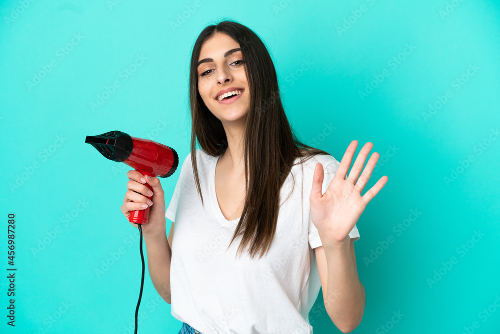 Young caucasian woman holding a hairdryer isolated on blue background saluting with hand with happy expression Young caucasian woman holding a hairdryer isolated on blue background saluting with hand with happy expression