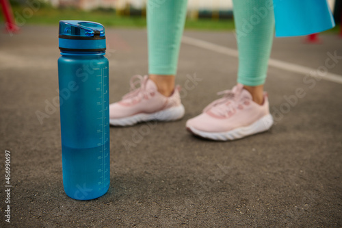 Close-up of a blue plastic bottle of water on the asphalt of sports ground next to sporty woman's feet. Health and body care concept