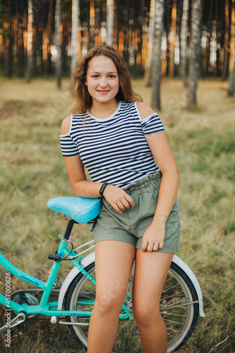 Portrait of a young girl with a blue bicycle. Girl in the woods on a bicycle. Striped T-shirt and dark green shorts. Cycling in the woods