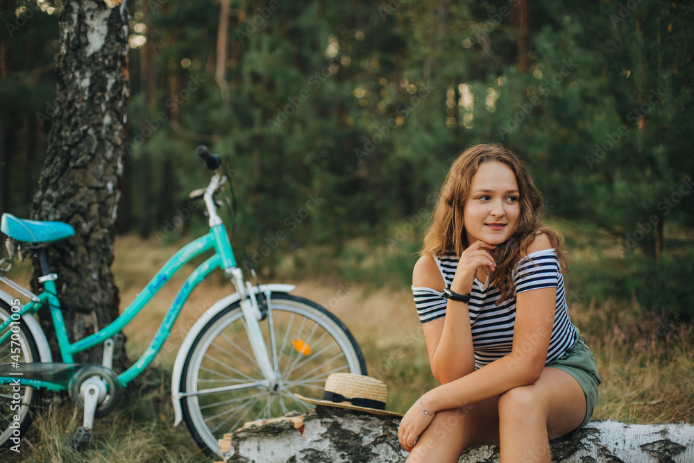 Beautiful young girl sitting on a fallen tree. Girl in a striped T-shirt and shorts. Portrait of a girl in the woods on the nature. Ride a bike in the woods Beautiful young girl sitting on a fallen tree. Girl in a striped T-shirt and shorts. Portrait of a girl in the woods on the nature. Ride a bike in the woods