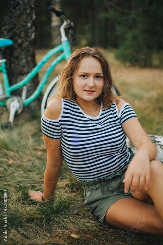 Beautiful young girl sitting on a fallen tree. Girl in a striped T-shirt and shorts. Portrait of a girl in the woods on the nature. Ride a bike in the woods