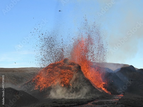 Volcanic vent at Fagradalsfjall, Iceland, erupting incandescent orange and red lava. Black cooled lava in the foreground and blue sky in the background. photo