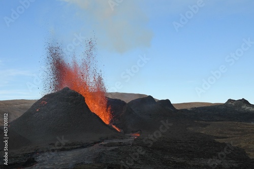 Volcanic vent at Fagradalsfjall, Iceland, erupting incandescent orange and red lava. Black cooled lava in the foreground and blue sky in the background.