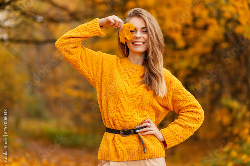 Happy beautiful young girl with a smile in a knitted yellow sweater covers her face with a yellow autumn leaf in a park with golden foliage. Colorful female style
