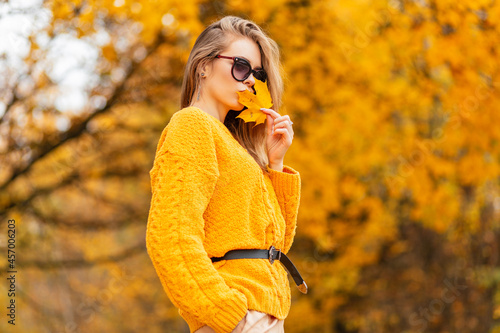 Fashionable beautiful young girl in a knitted yellow sweater kisses an autumn golden leaf in a park with bright orange foliage