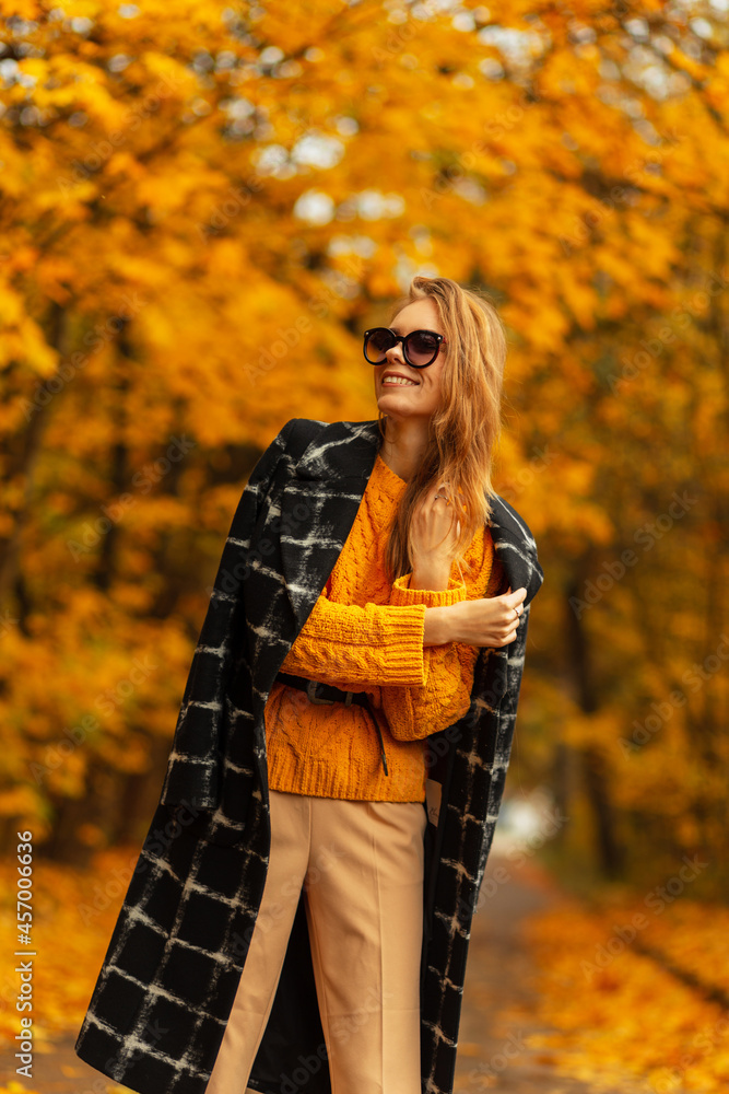 Happy pretty young woman with beautiful smile with sunglasses in trendy black coat with vintage yellow sweater enjoying autumn weather outdoors with colorful yellow foliage