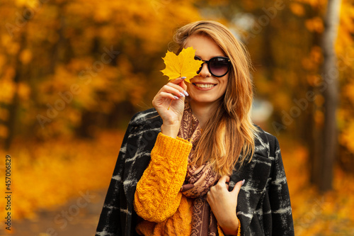 Happy beautiful young girl with a sweet smile in a yellow vintage knitted sweater and a fashionable black coat with sunglasses covers face with a yellow leaf in amazing nature with colorful leaves
