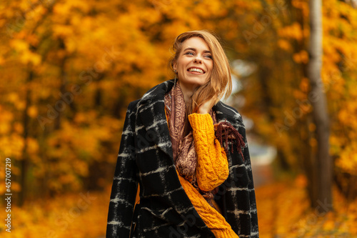Happy beautiful young girl with a cute smile in a fashionable coat with a knitted yellow sweater and a scarf walks outdoors with autumn trees and bright yellow foliage. Expression of positive emotion