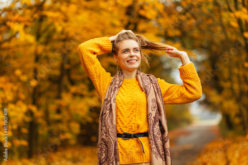 Happy emotional girl with a cute smile in a fashion vintage sweater and scarf is walking in the park with colored autumn yellow foliage. Positive female face