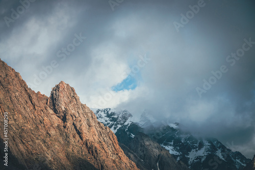 Awesome landscape with sunlit rocky pinnacle on background of high snowy mountains in low clouds. Atmospheric alpine scenery with sharp rock and giant snowy mountain in cloudy sky. Scenic alpine view.