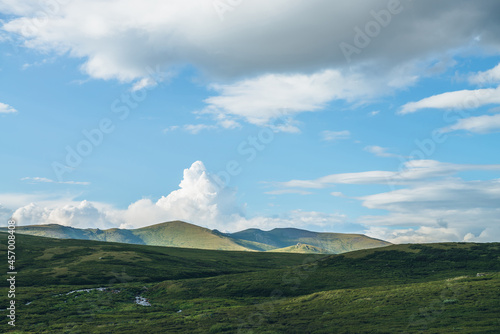 Green sunny mountain landscape with big white cloud in form of explosion in blue sky above green hills in sunlight. Beautiful sunny scenery with big cloud in shape of explosion above green mountains.