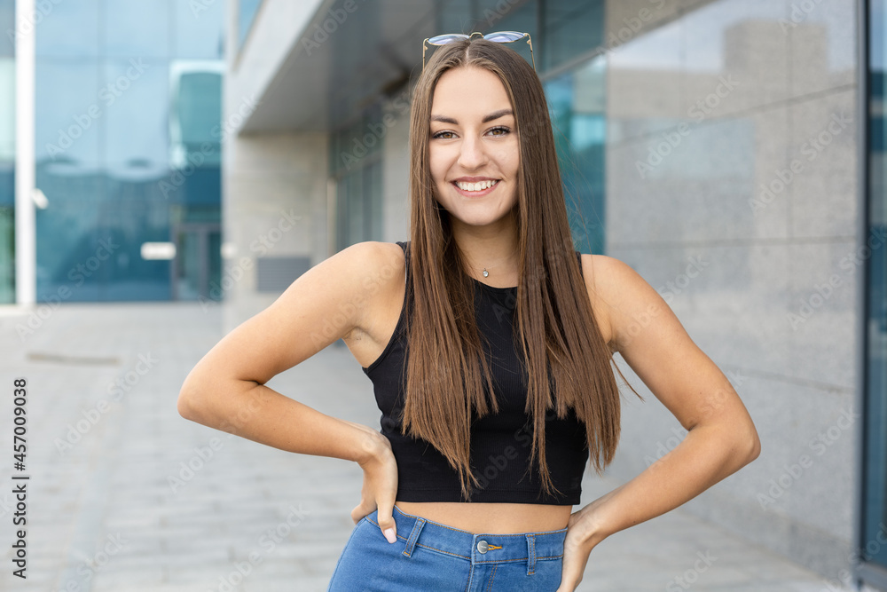 Lifestyle, femininity, body and people concept - A beautiful girl with straight dark hair and glasses on head wearing a black top and jeans standing on the street with one arm around her waist