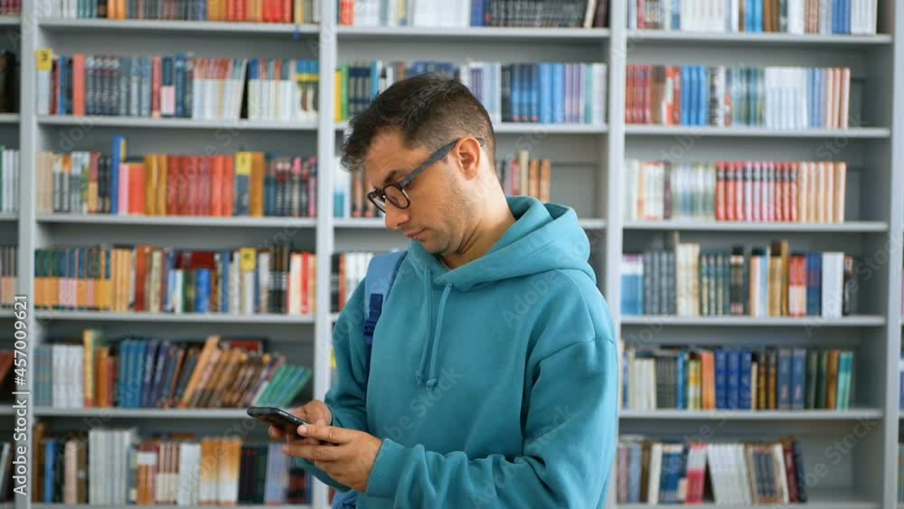 Close-up portrait of a young attractive student guy, he is texting with friends on his smartphone against the background of bookshelves in the university library. Close-up portrait of a young attractive student guy, he is texting with friends on his smartphone against the background of bookshelves in the university library.
