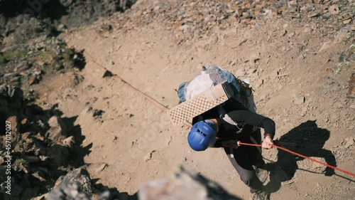 The climber holds on to the rope and descends a difficult section in the mountains. A man with a large backpack with equipment descends with a rope in the mountains. View from above. Close-up photo