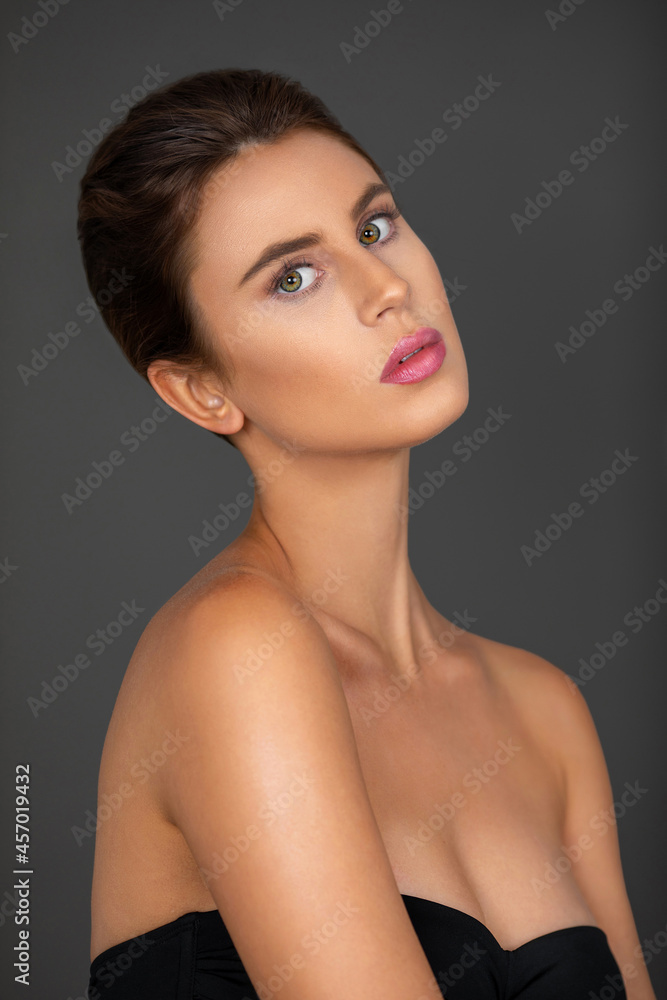 Naklejka premium Portrait of a sexy young white woman with wavy hair in a bun and beautiful makeup and lipstick posing by herself inside a studio with a grey background.