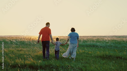 Dad, son, mom, play together run, happy child holds hands of parents, child is jumping on green grass. Family walk in park in spring at sunset, healthy childhood. Family weekend in summer in nature